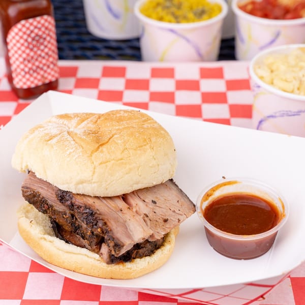 Larry J's BBQ Cafe brisket sandwich with a side of BBQ sauce and various sides in the background.